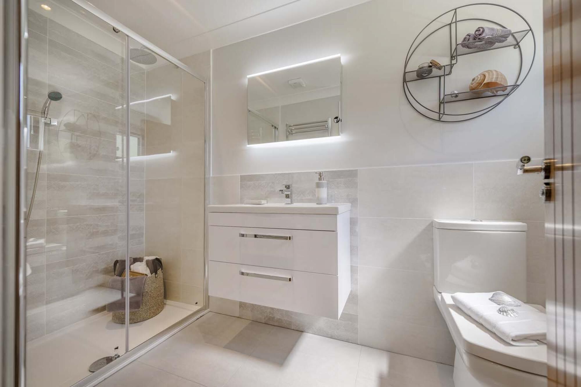 Une salle de bain moderne avec une douche à l’italienne en verre, un lavabo blanc avec un miroir au-dessus, et des toilettes sur lesquelles est posée une serviette.