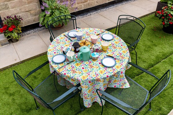 A round table with a floral tablecloth, surrounded by four chairs, set on a patch of artificial grass.