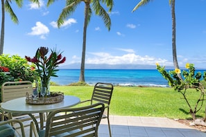 A patio with a table and chairs overlooking a beach with palm trees and a clear blue sky. - Napili Bay beauty, a cozy oceanfront studio (Lahaina)