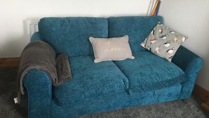 A teal sofa with a 'Love' pillow and a bird-patterned pillow, a brown blanket, and a flower decoration on the wall. - Driftwood Loft (Downpatrick)
