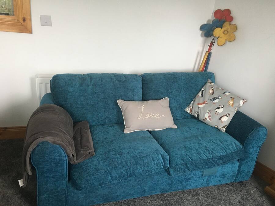 A teal sofa with a 'Love' pillow and a bird-patterned pillow, a brown blanket, and a flower decoration on the wall.