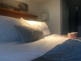 Uma cama bem arrumada com travesseiros, uma mesa de cabeceira com xícara e pires, e um quadro na parede.