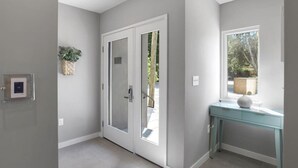 Un couloir avec une porte donnant sur un balcon, une fenêtre offrant une vue sur la verdure et une petite table avec une lampe.