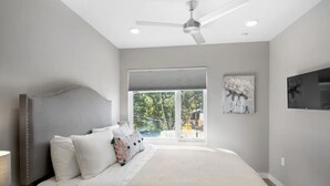 Une chambre avec un grand lit, un ventilateur de plafond, une fenêtre avec des stores et un téléviseur fixé au mur.
