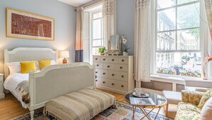 Une chambre à coucher comprenant un lit, un canapé, une commode, un miroir et une fenêtre donnant sur des immeubles.