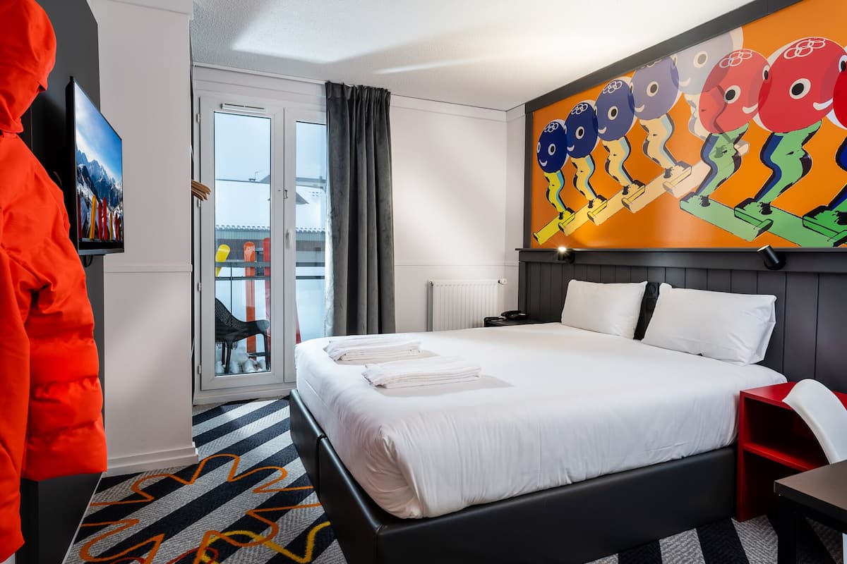 A hotel room with a bed, a television, a painting, and a view of the outside.