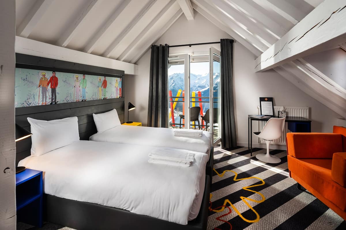A hotel room with a large bed, a desk with a chair, a view of mountains through a glass door, and a colorful artwork on the wall.