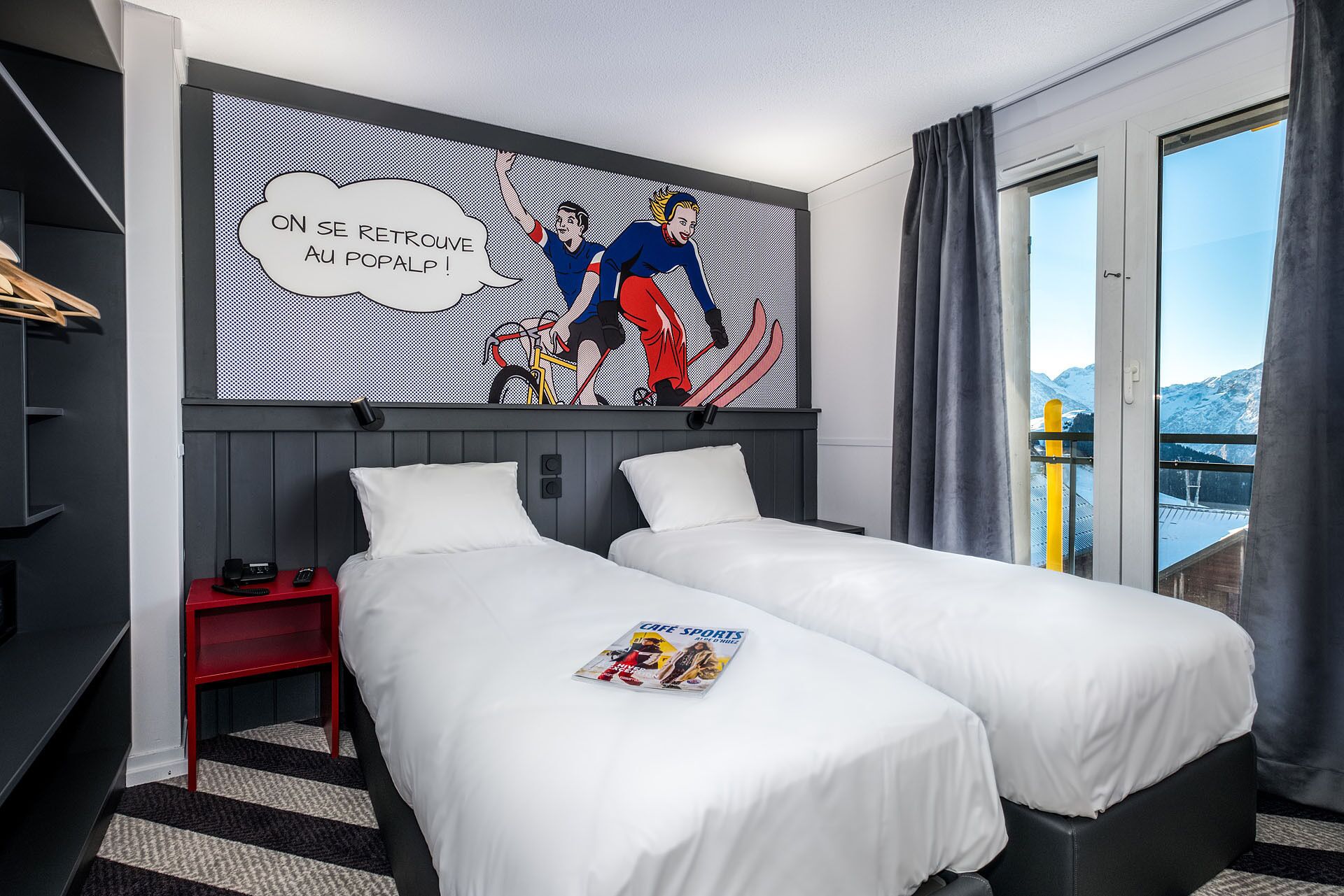 A hotel room with two beds, a red nightstand, a magazine on the left bed, and a large window with a view of mountains.