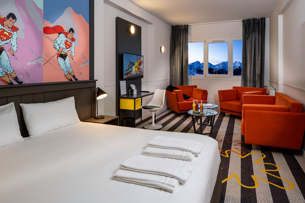 A hotel room with a large bed, two orange armchairs, a small table, and a painting on the wall.