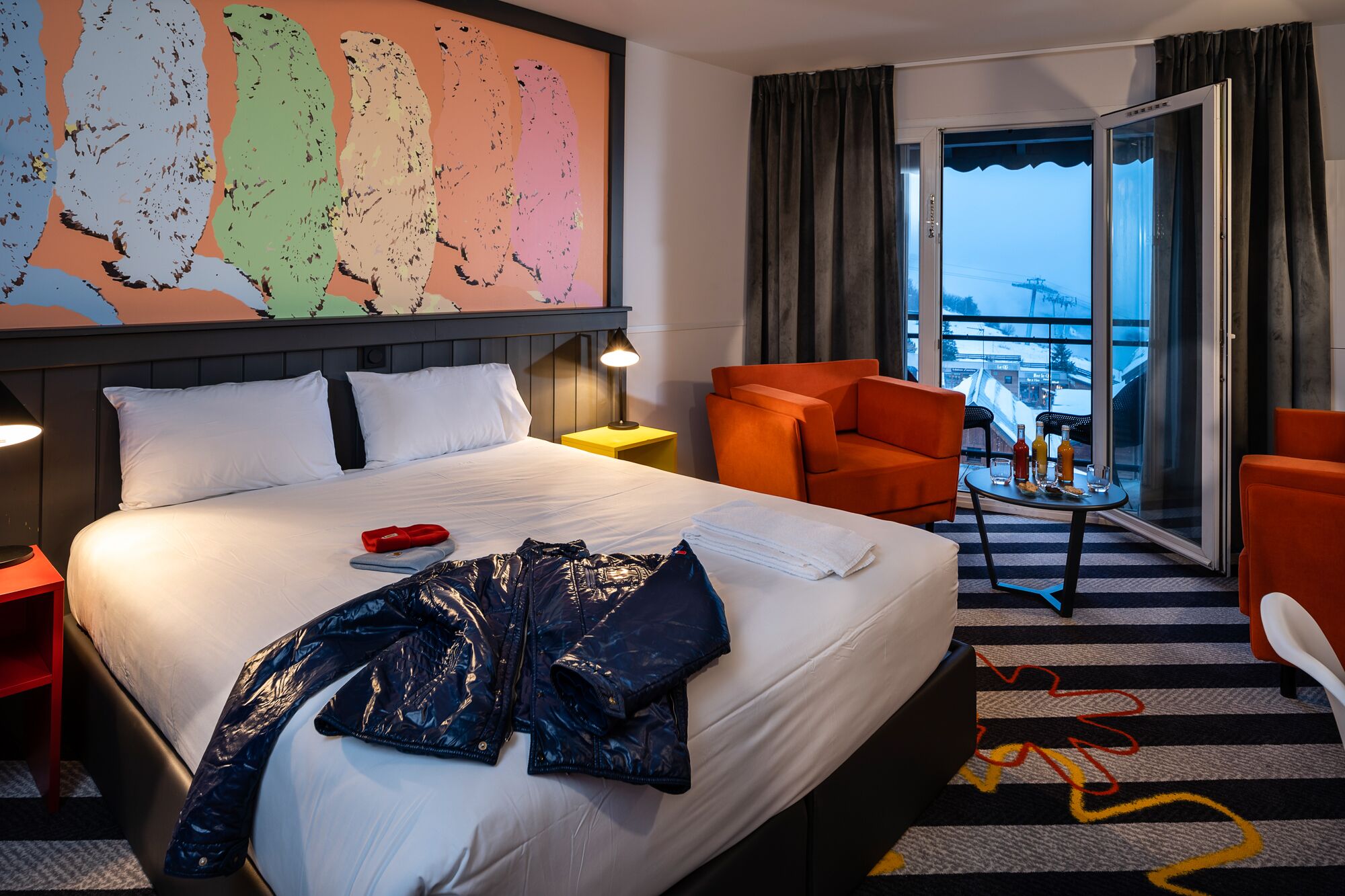Un cuarto de hotel con una cama grande, un escritorio con una lámpara, un sillón naranja y vistas a un paisaje nevado a través de la ventana.