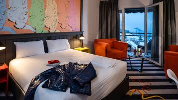 Un cuarto de hotel con una cama grande, un escritorio con una lámpara, un sillón naranja y vistas a un paisaje nevado a través de la ventana.