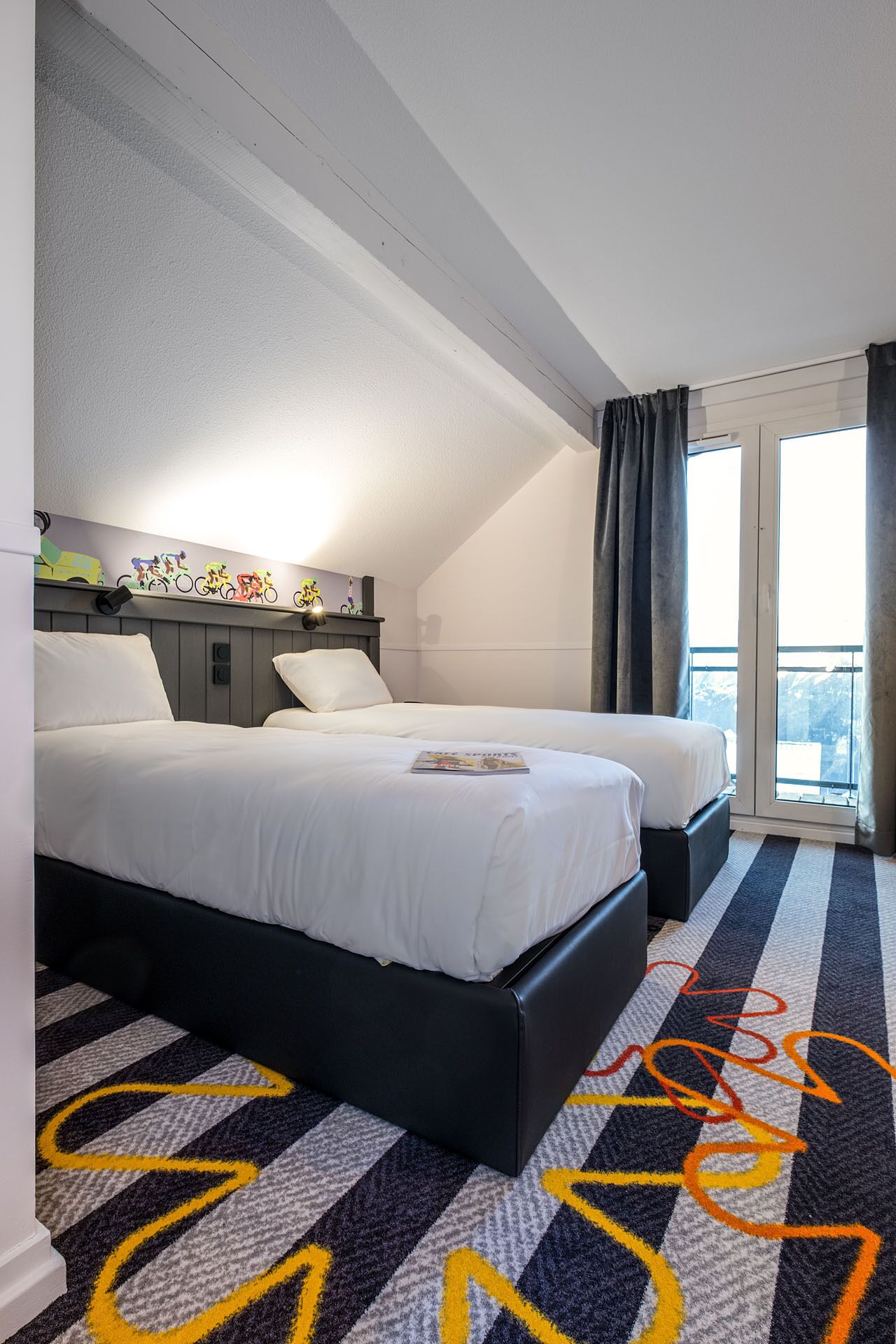Habitación de hotel con dos camas, un ventanal con cortinas, una lámpara de pared y un cuadro decorativo de una bicicleta.