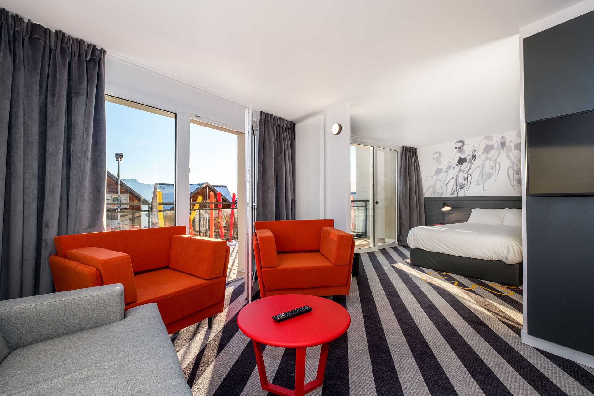 Ein Hotelzimmer mit einem Bett, zwei orangenen Sofas, einem roten Tisch und einem großen Fenster mit Vorhängen.