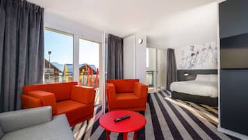 Ein Hotelzimmer mit einem Bett, zwei orangenen Sofas, einem roten Tisch und einem großen Fenster mit Vorhängen.