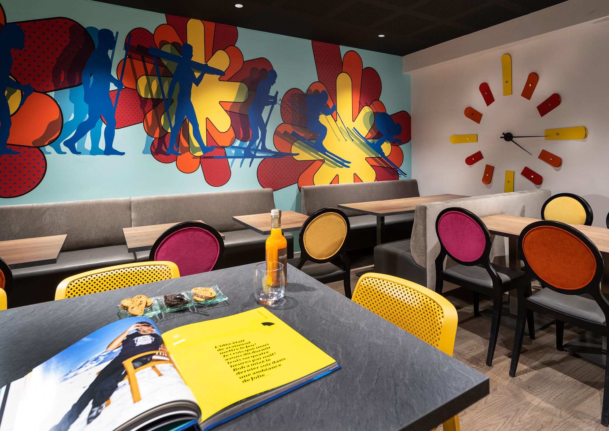 Un interior de restaurante moderno con arte colorido en las paredes, un reloj y diversas opciones de asientos, incluyendo mesas y cabinas.