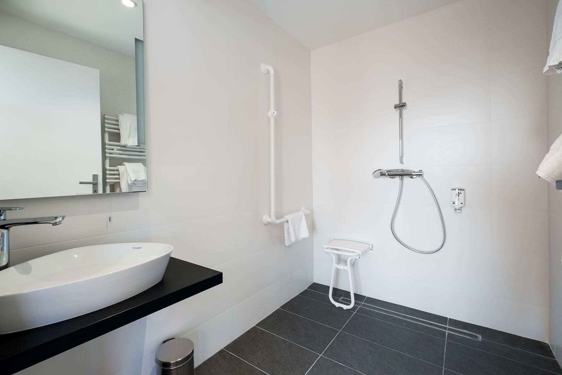 A modern bathroom with a large white sink, a mirror, a shower with a handheld showerhead, and a towel rack.
