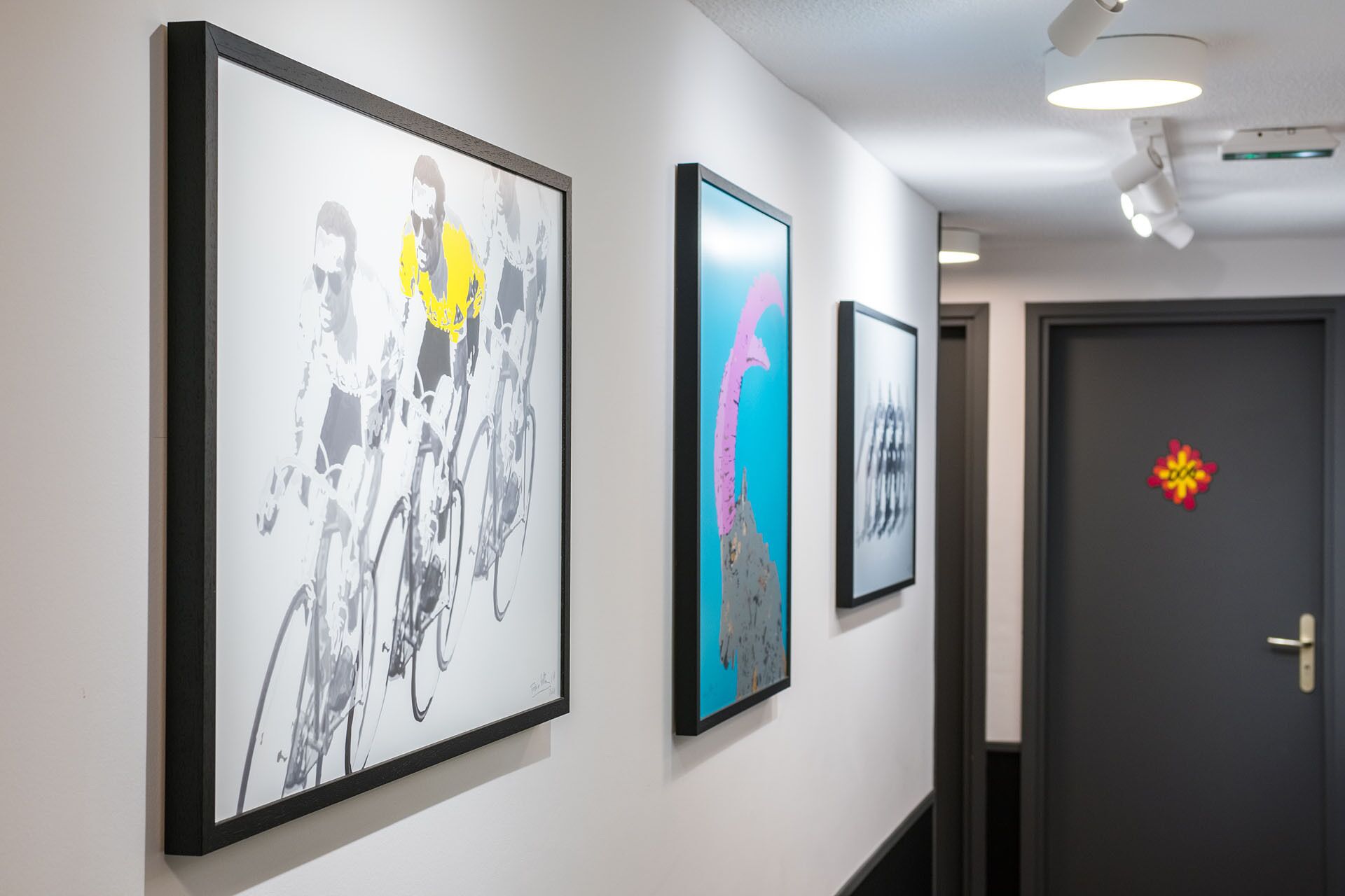 A hallway with three framed artworks on the wall, a door with a decorative sticker, and recessed ceiling lights.