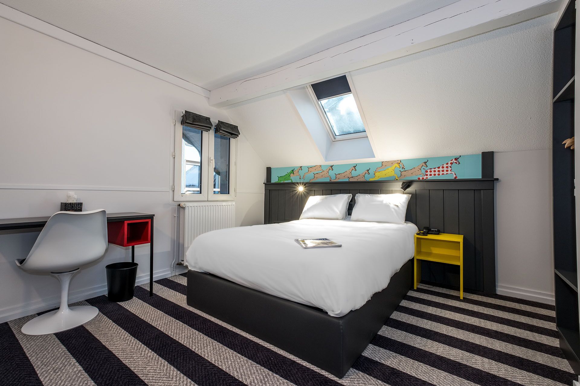 A hotel room with a bed, a chair, a small table, a radiator, and a window with a view.