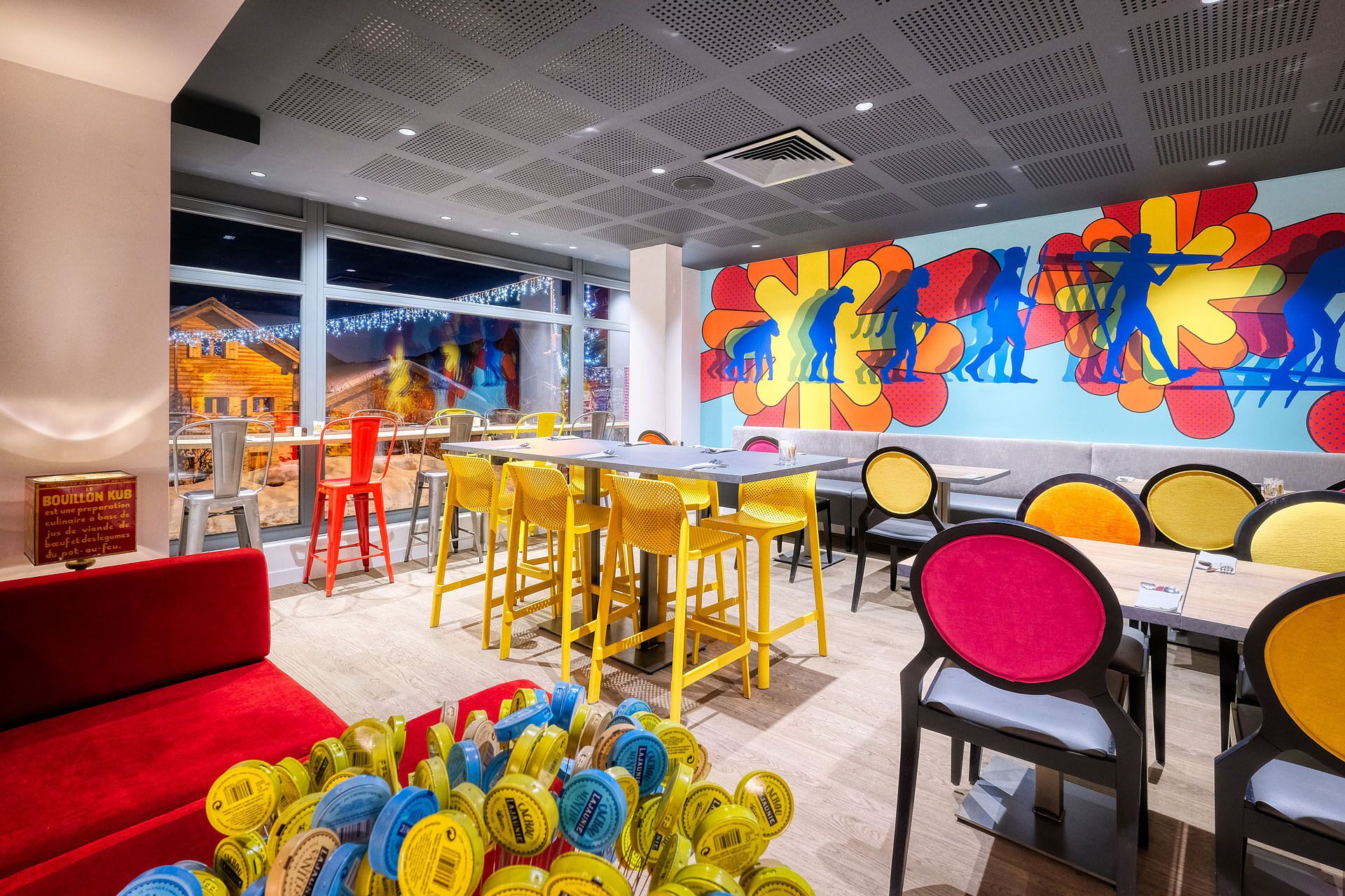 Un restaurante moderno con sillas amarillas y rojas, un mural colorido y vistas a un pueblo nevado a través de amplios ventanales.