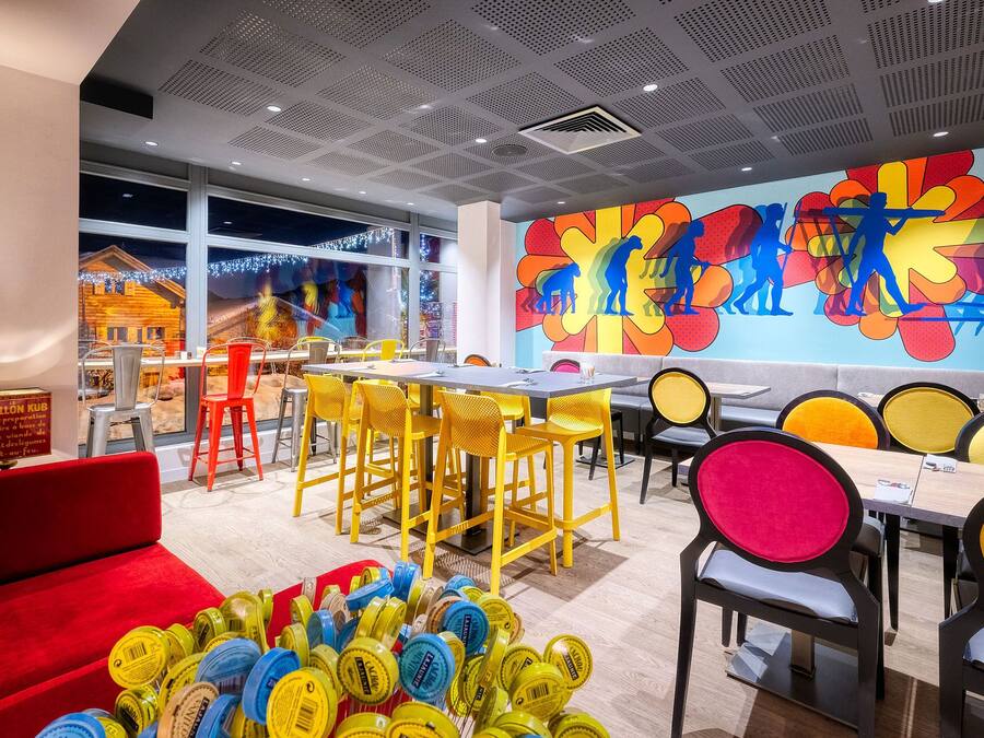 Un restaurante moderno con sillas amarillas y rojas, un mural colorido y vistas a un pueblo nevado a través de amplios ventanales.