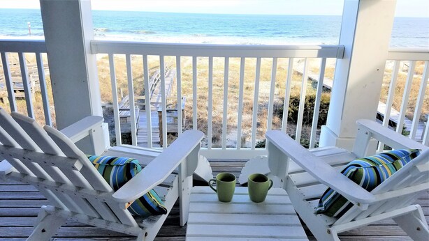 Una terraza de madera con dos sillas blancas y dos tazas verdes, con vista a una playa que tiene pasarelas de madera.