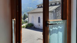 Une vue à travers une fenêtre ouverte, donnant sur un bâtiment à la façade en pierre, une cour intérieure et des montagnes en arrière-plan.