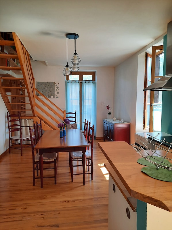Une cuisine spacieuse avec un plancher en bois, un coin repas avec une table et des chaises en bois, un escalier et une vue sur l’extérieur par une fenêtre.