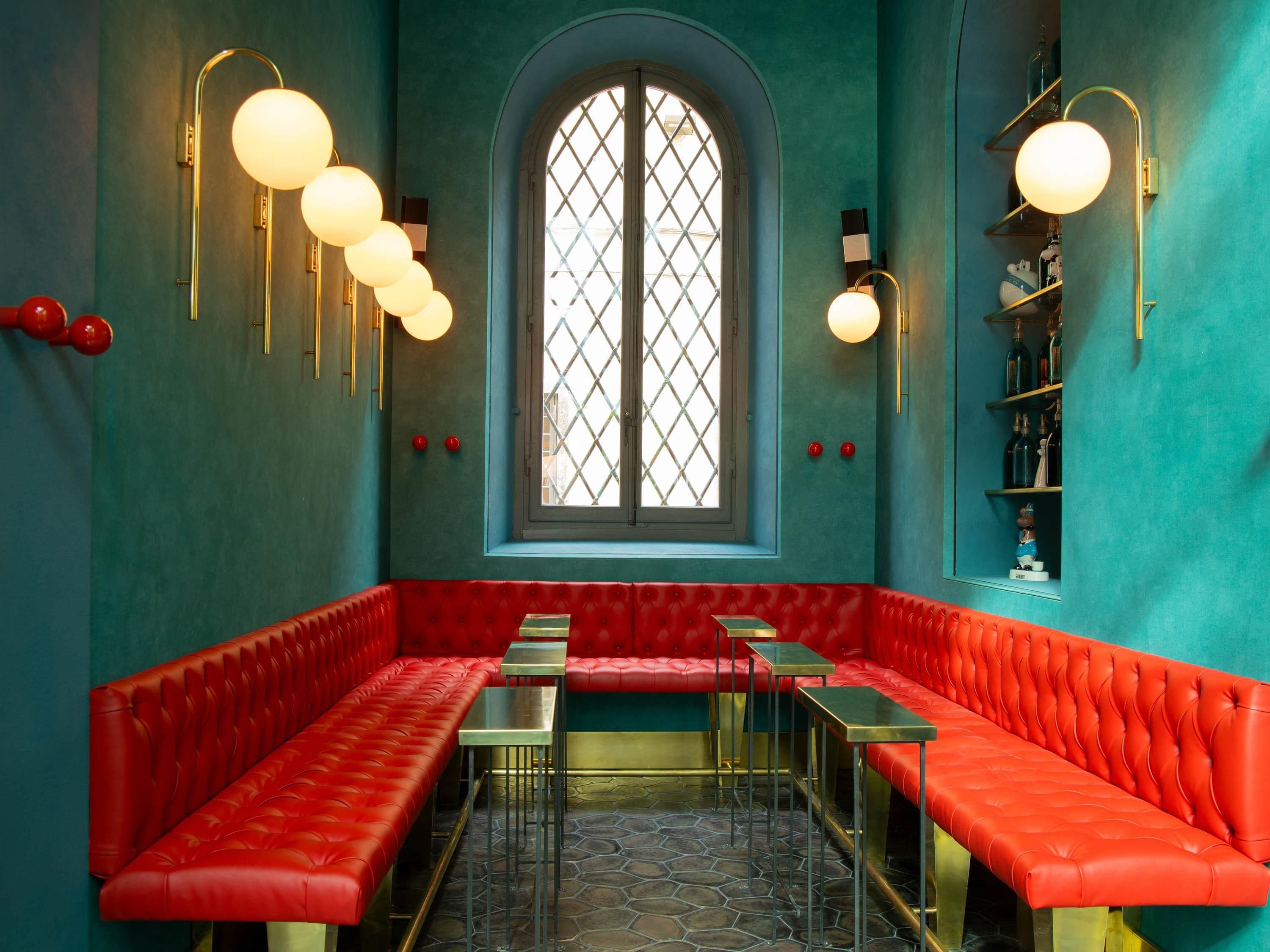 Een zithoek met rode, gestoffeerde banken, een groot rondboogvenster en bolvormige lampen aan de muur.