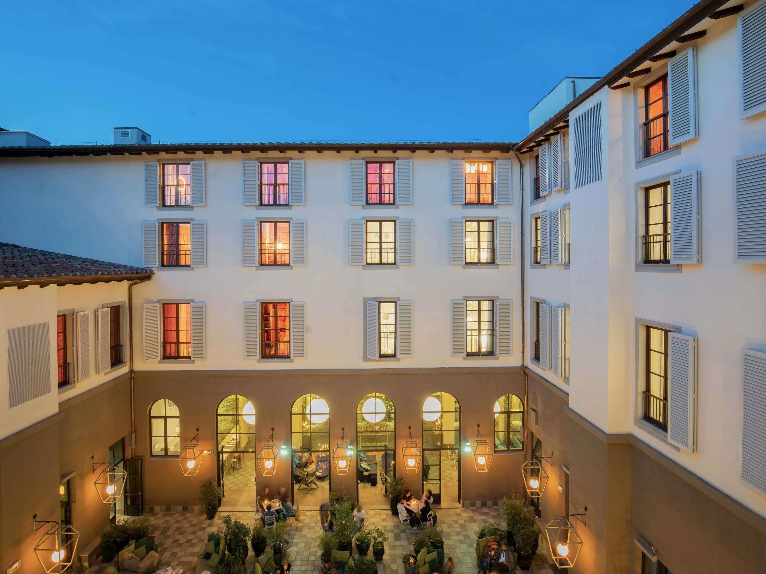 A courtyard with multiple seating areas and arched windows, surrounded by buildings with multiple windows and shutters.