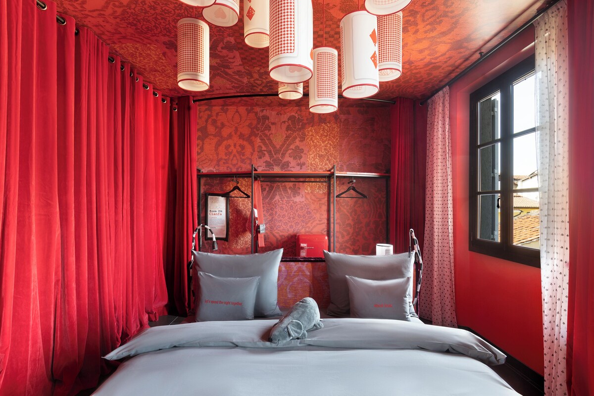 A hotel room with red curtains, a patterned wall, a bed with pillows, and a window with curtains.