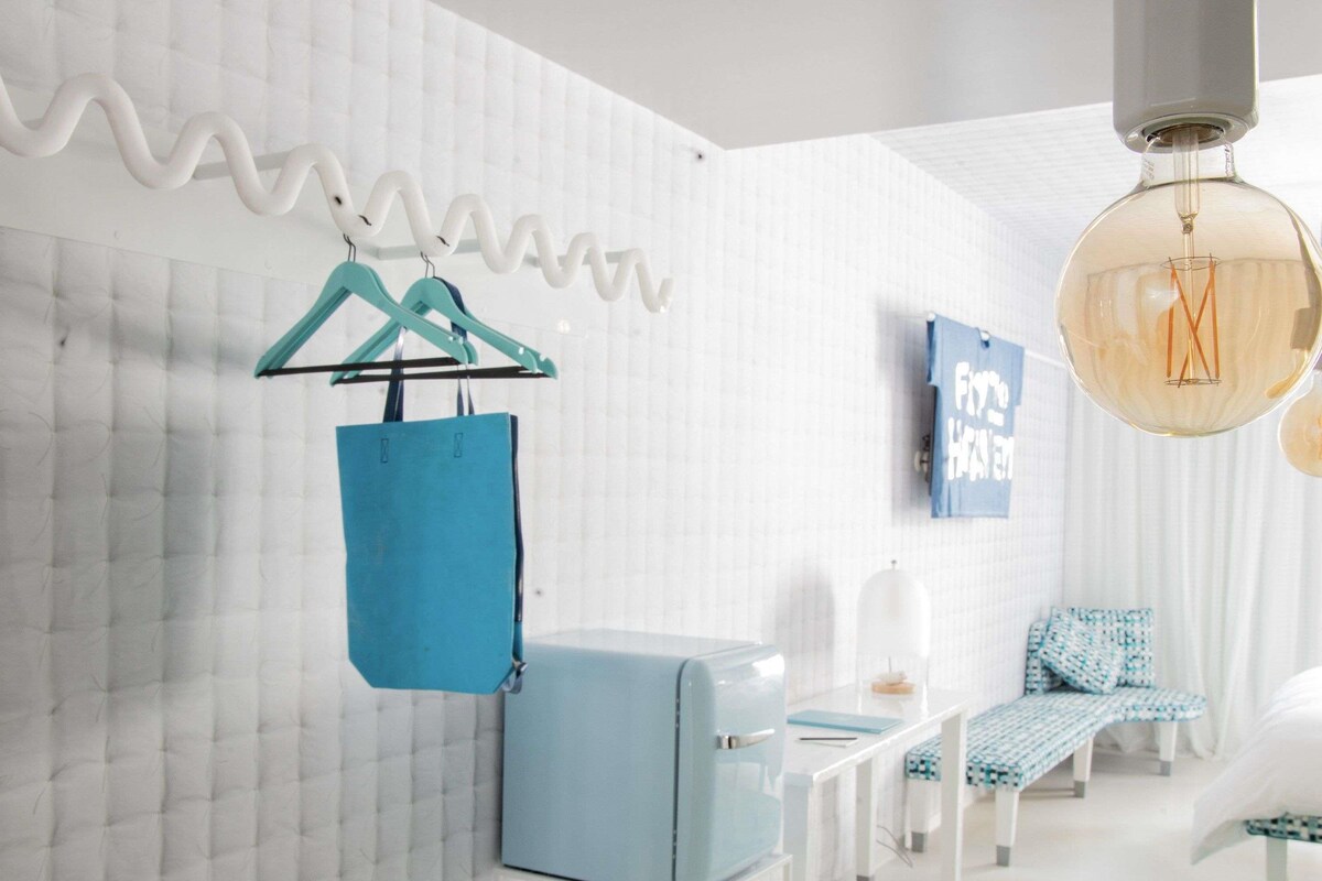 A modern hotel room with a white textured wall, a blue suitcase, a white chair, a blue sofa, and a hanging light fixture.