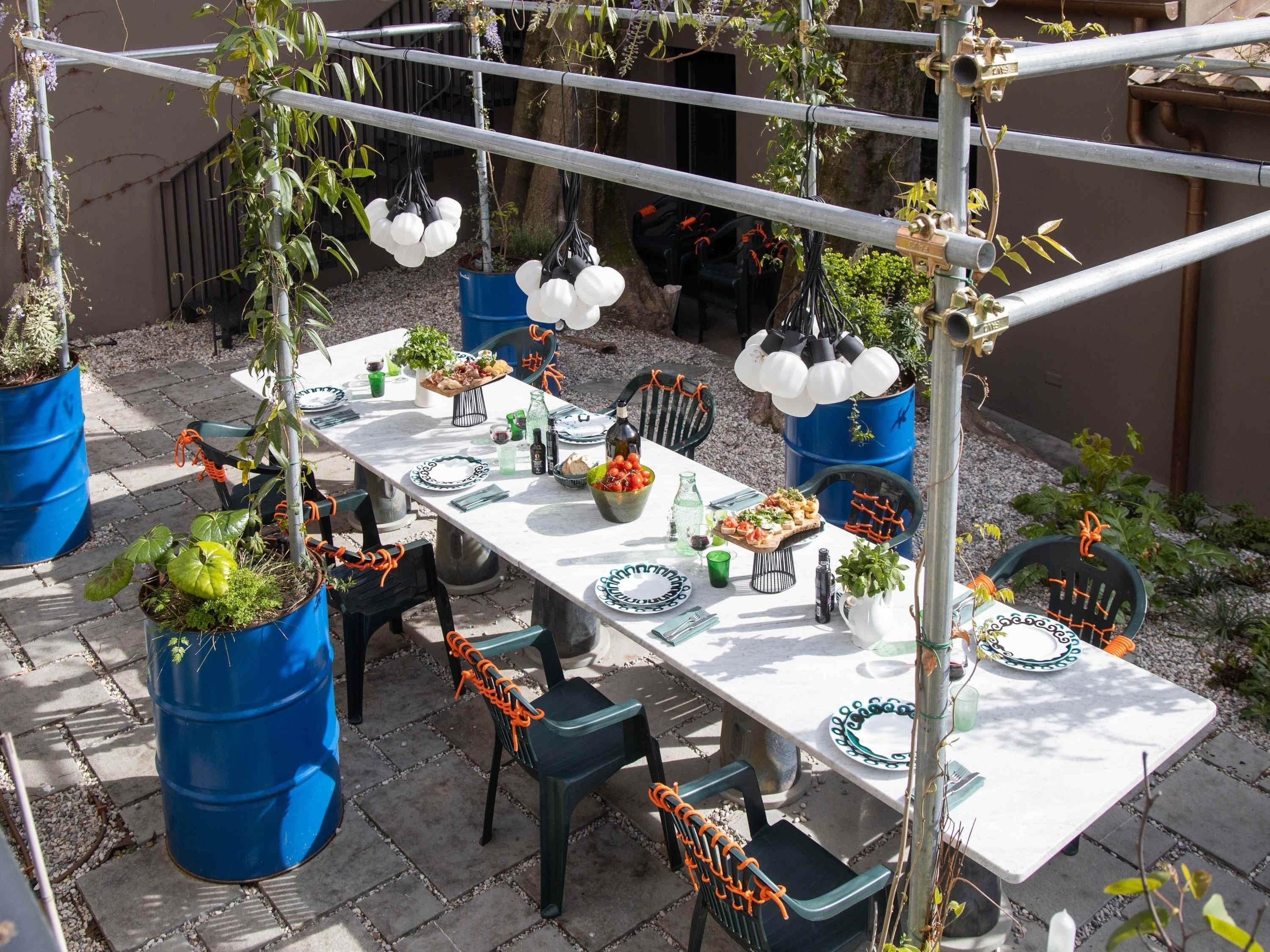 An outdoor dining area with a long table set for a meal, surrounded by chairs and potted plants.