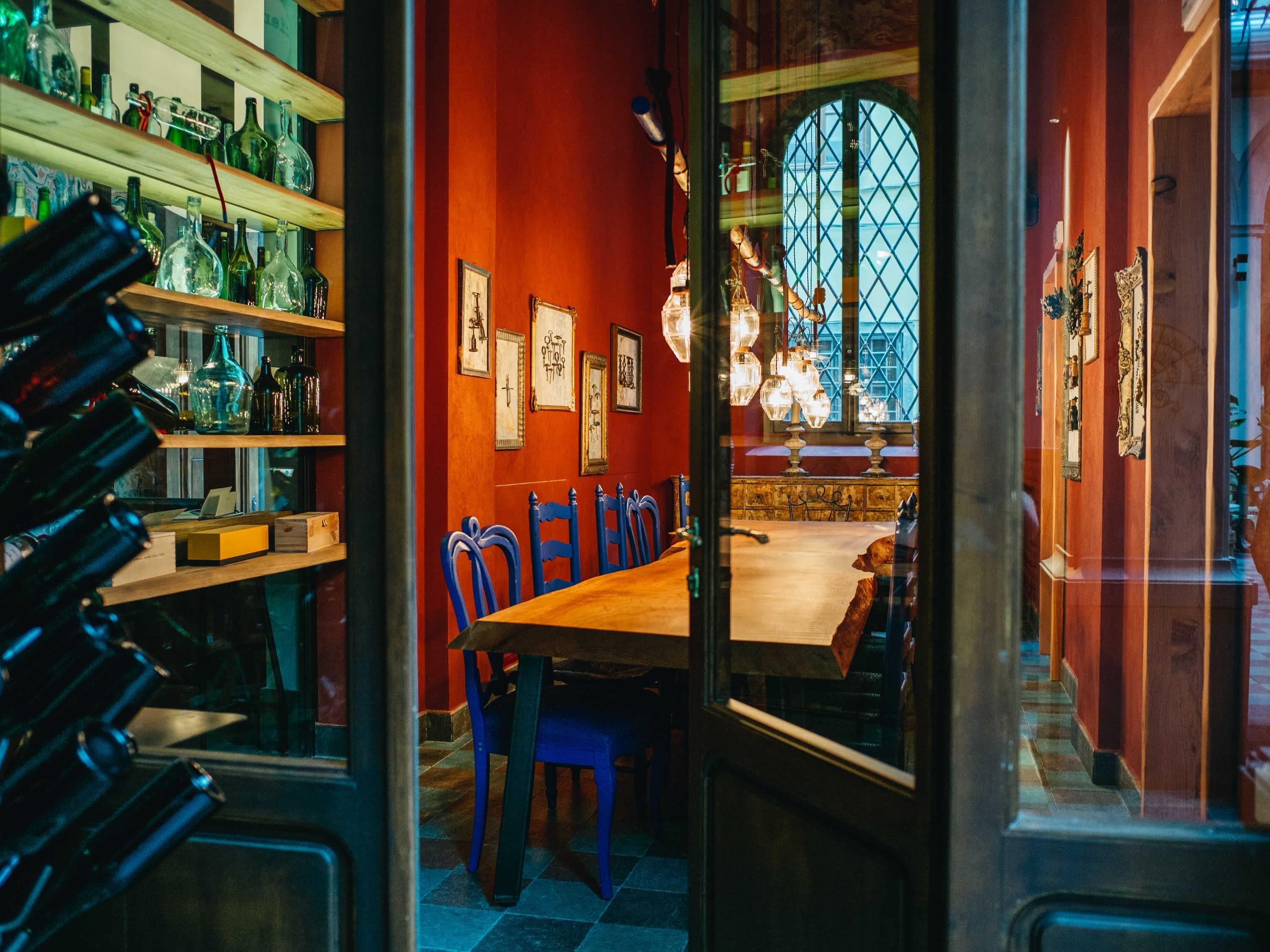 A dining area with a wooden table and blue chairs, shelves with bottles, and a large arched window with a view.