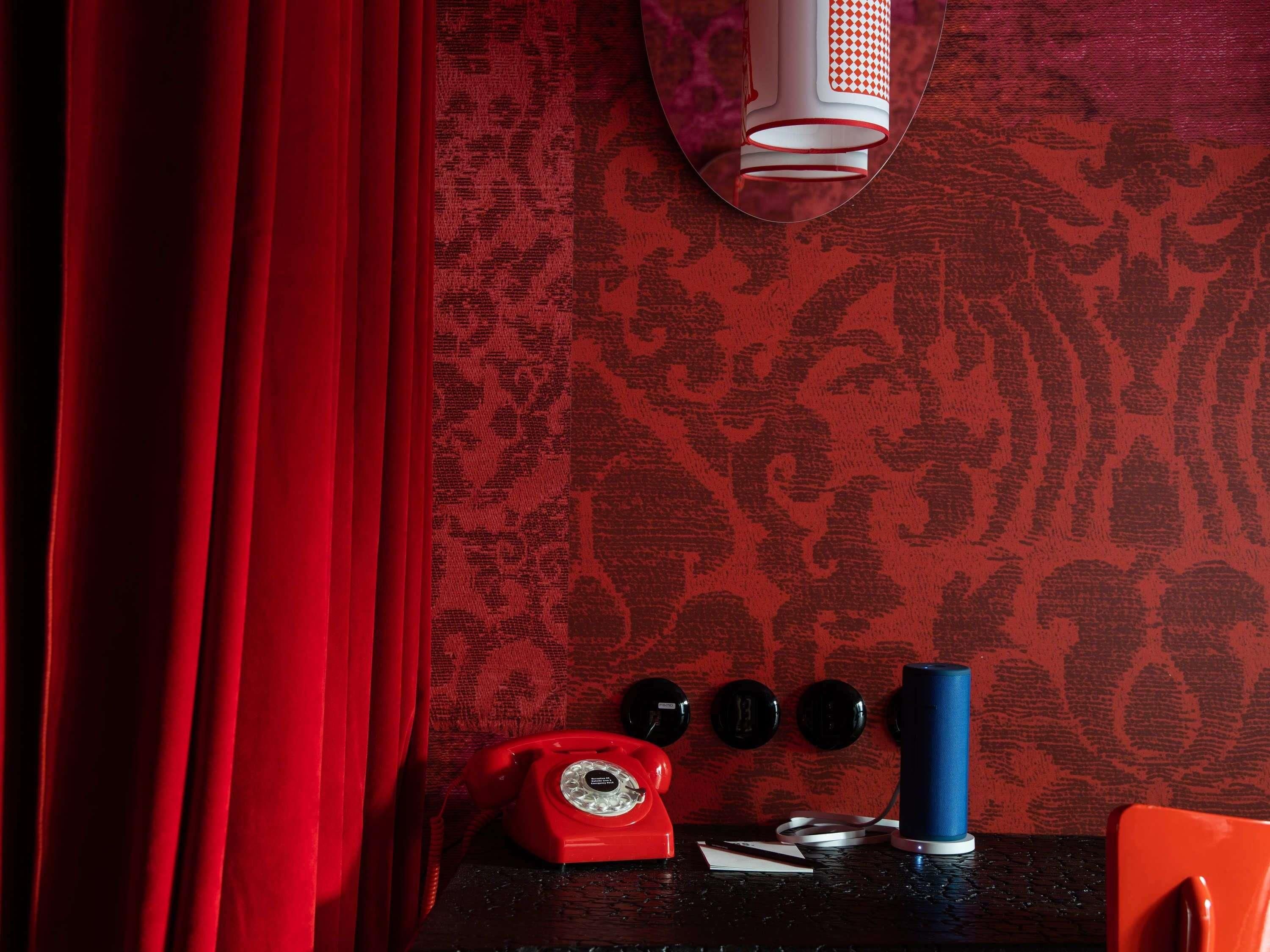 A room with a red curtain, a patterned wallpaper, a red rotary phone, a blue cylindrical object, and black circular objects on a dark surface.