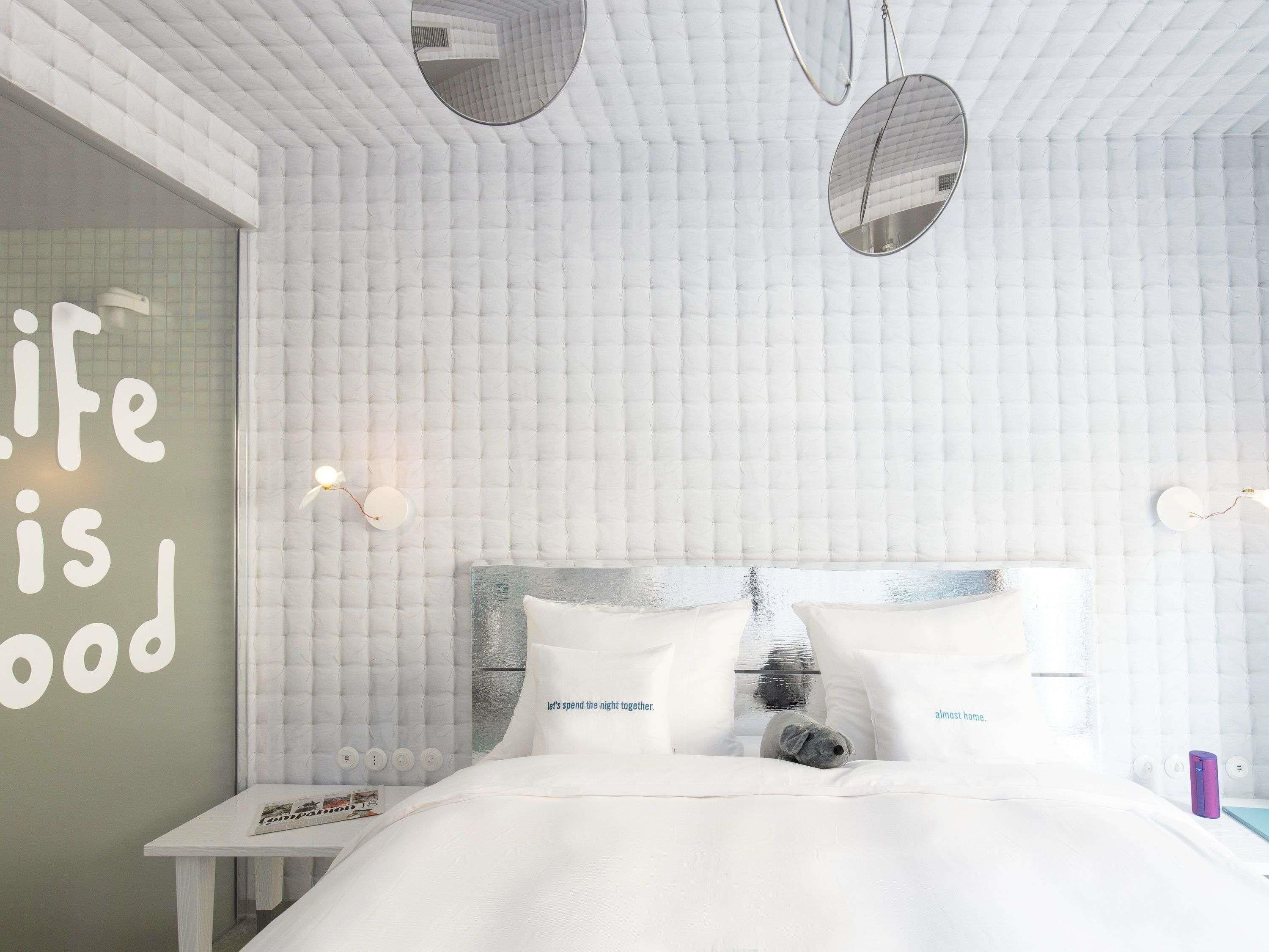 A bedroom with a tufted wall, a bed with white bedding, a small table with a lamp, and a wall with a motivational quote.