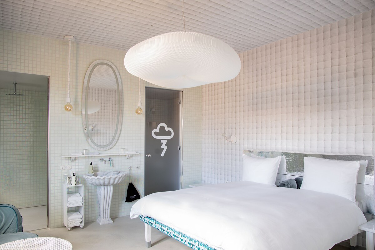 A modern bedroom with a large bed, a round mirror, a white sink, and a door with a weather symbol.