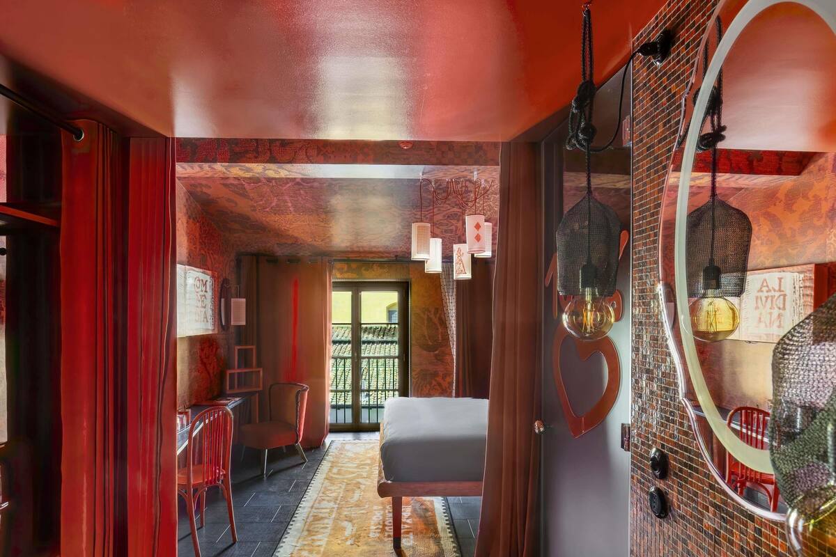 Une pièce comprenant un lit, un miroir, une chaise rouge, une table et un tapis à motifs.