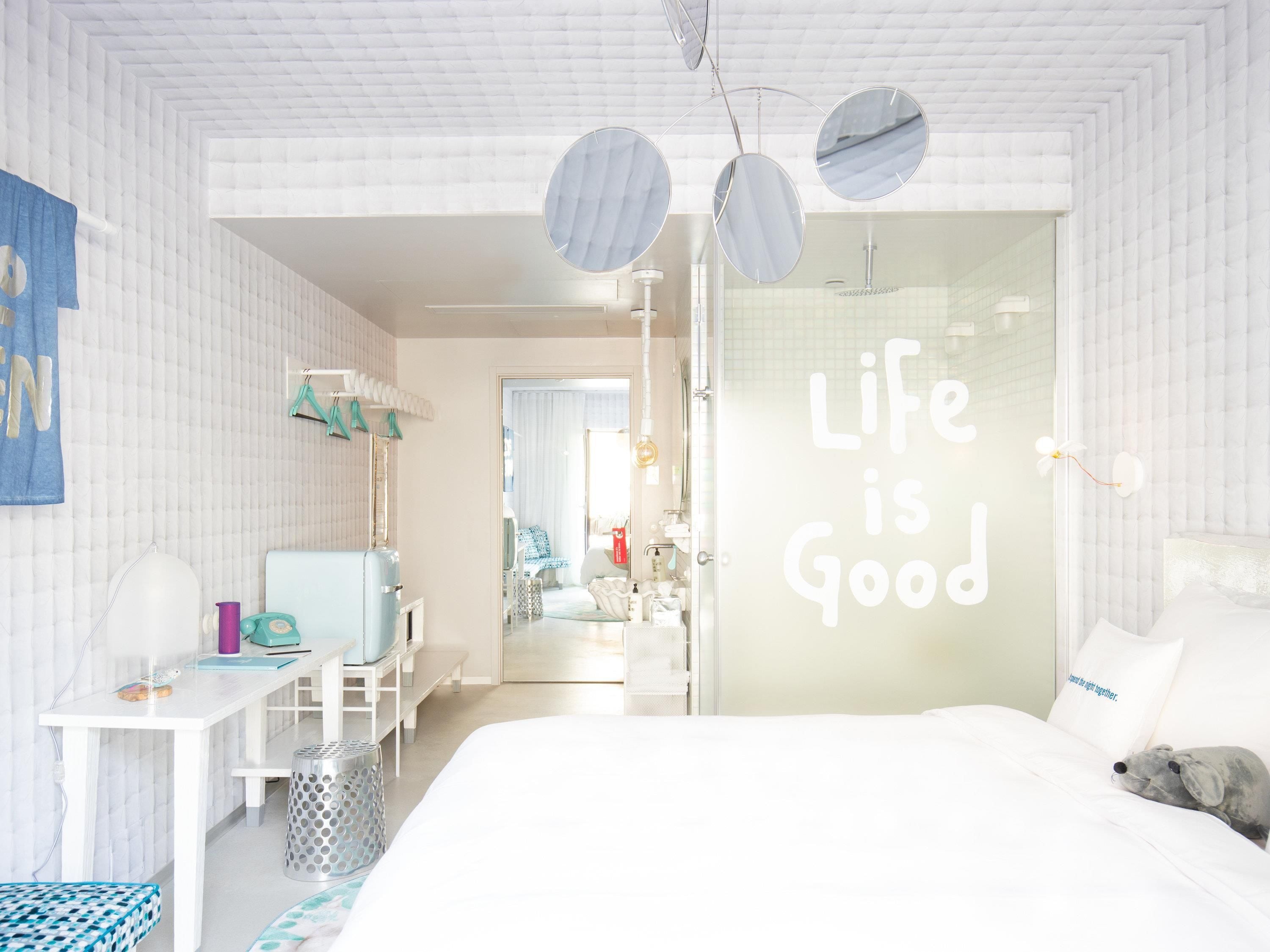 A modern bedroom with a bed, a desk, and a mirror. There's a message on the wall that says 'Life is Good'.