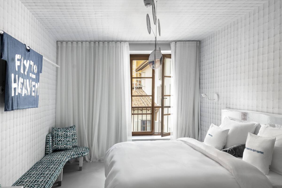 Une chambre d’hôtel avec un lit, un banc, une fenêtre donnant sur un paysage agréable et un panneau fixé au mur.
