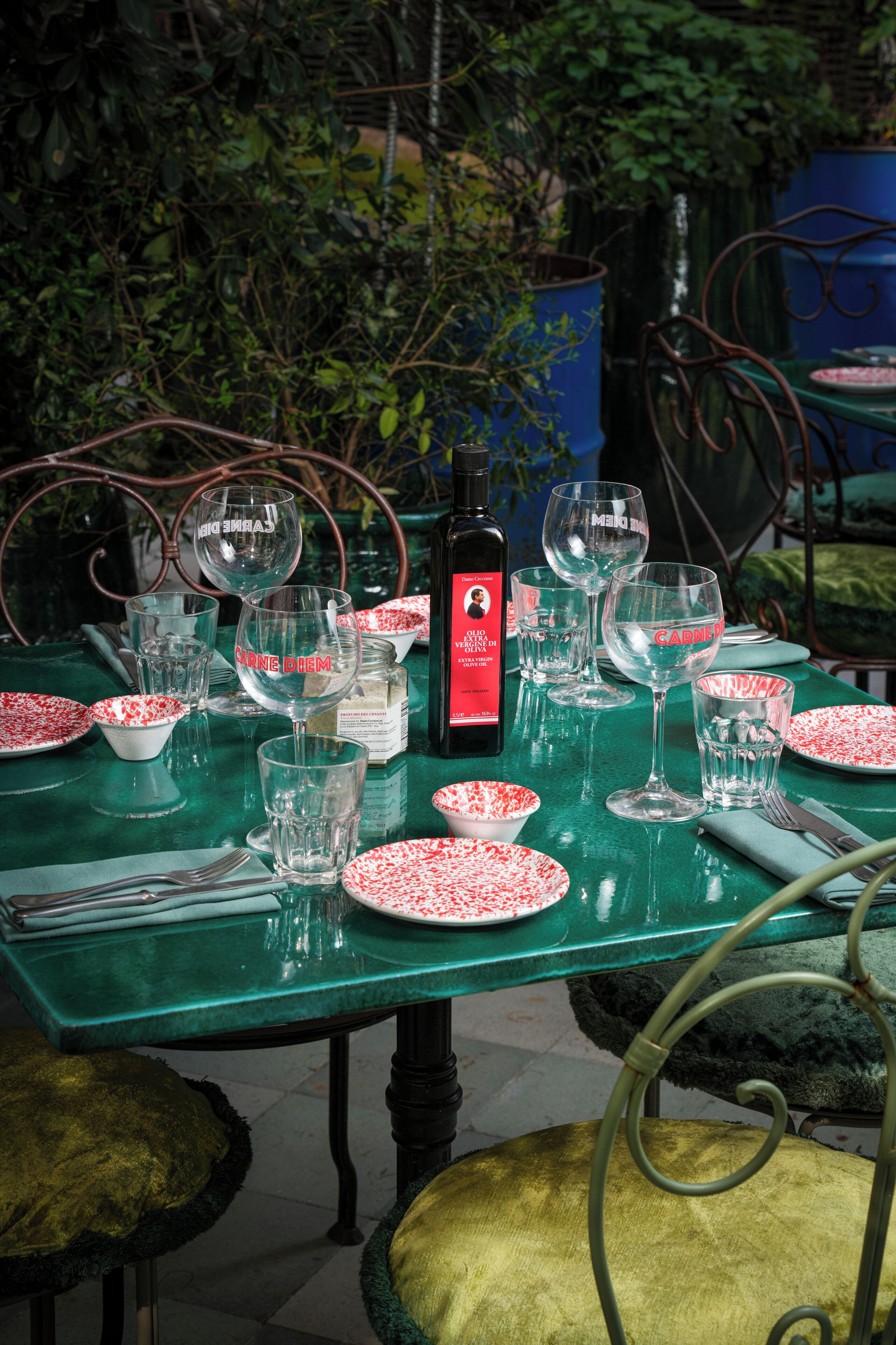 A table set with wine glasses, plates, and a bottle of wine, surrounded by chairs and greenery.
