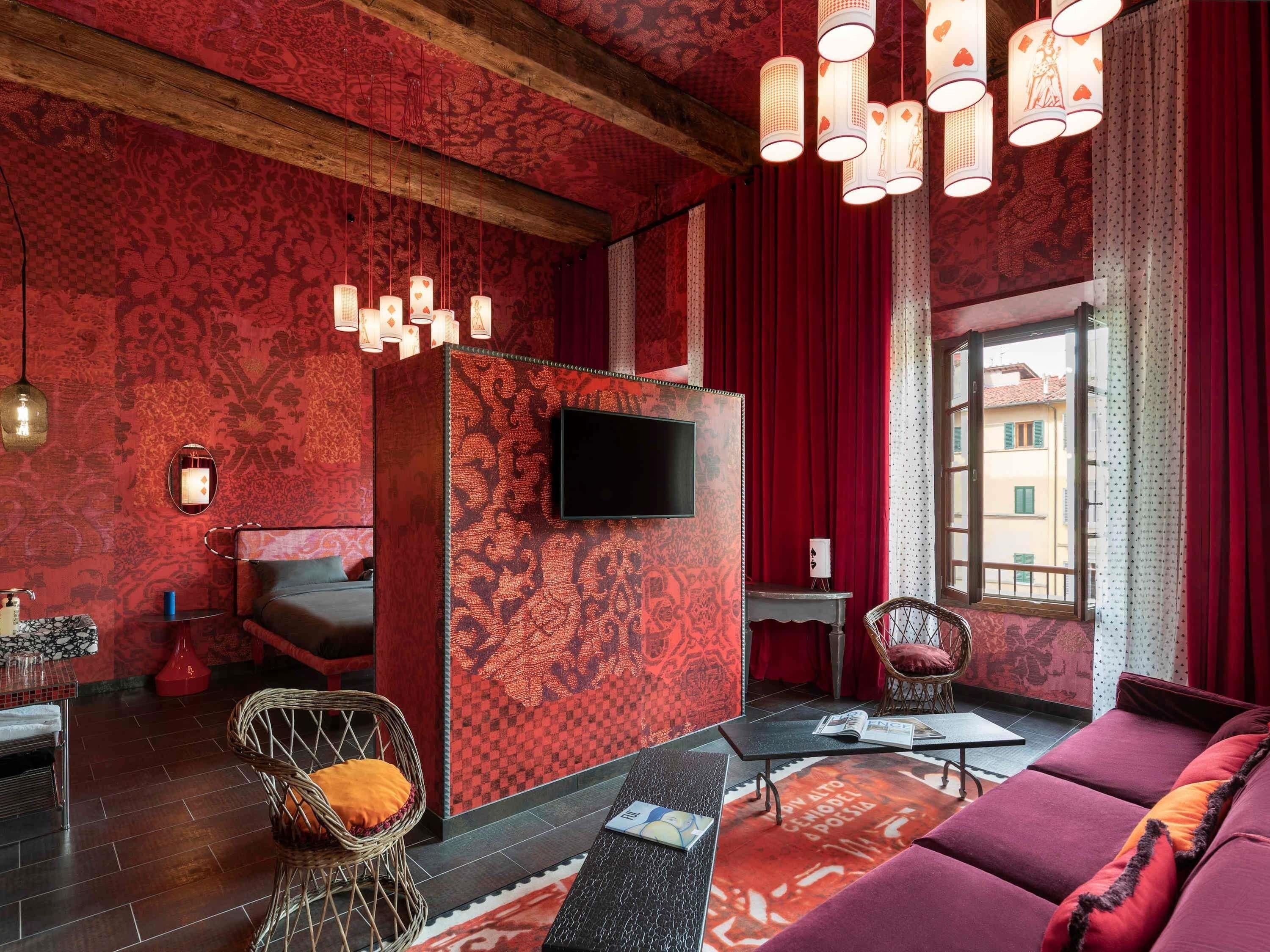 A room with a red patterned wall, a flat-screen TV, a sofa, a wicker chair, and a small table with a magazine.