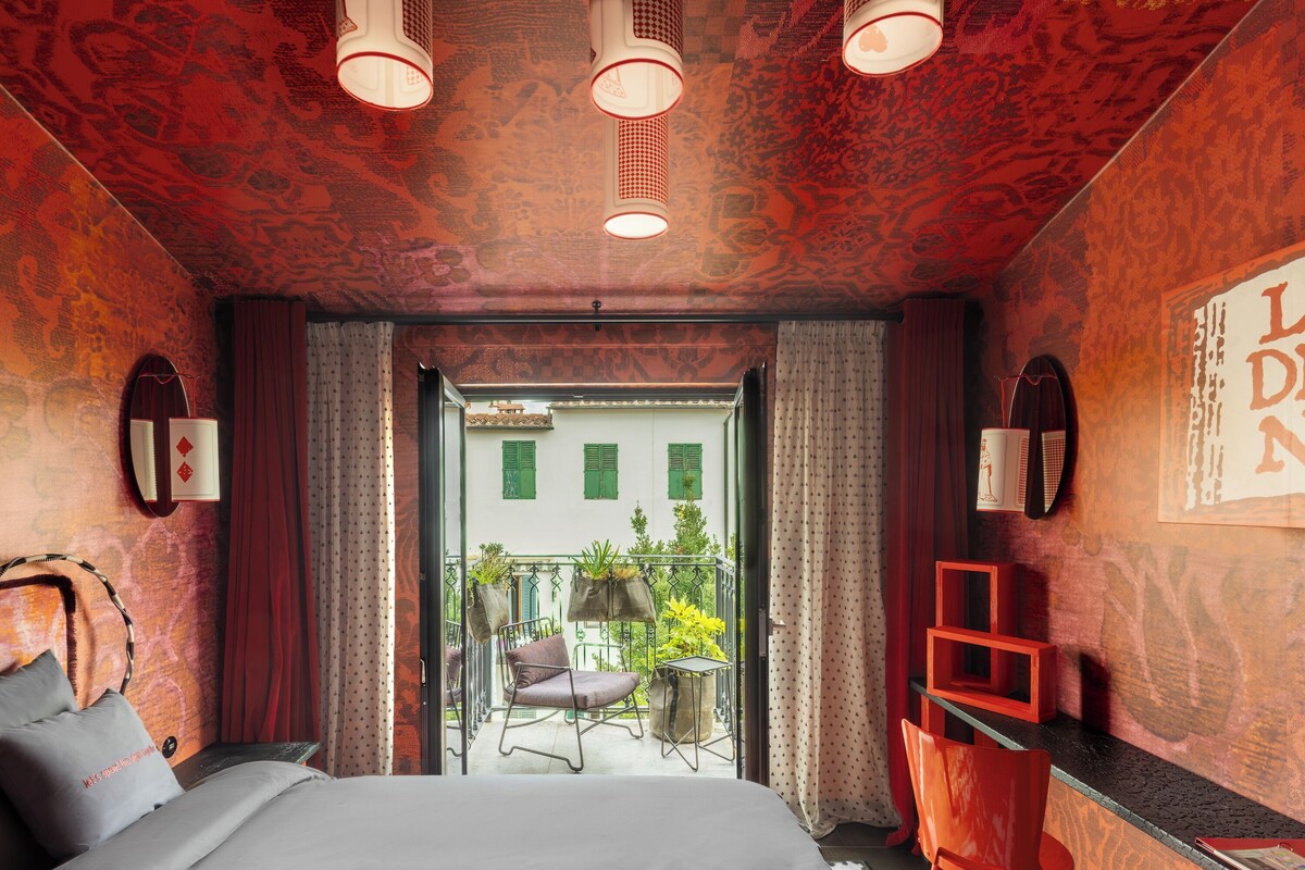 A hotel room with a bed, a chair, a small table, a red shelf, and a balcony with plants.