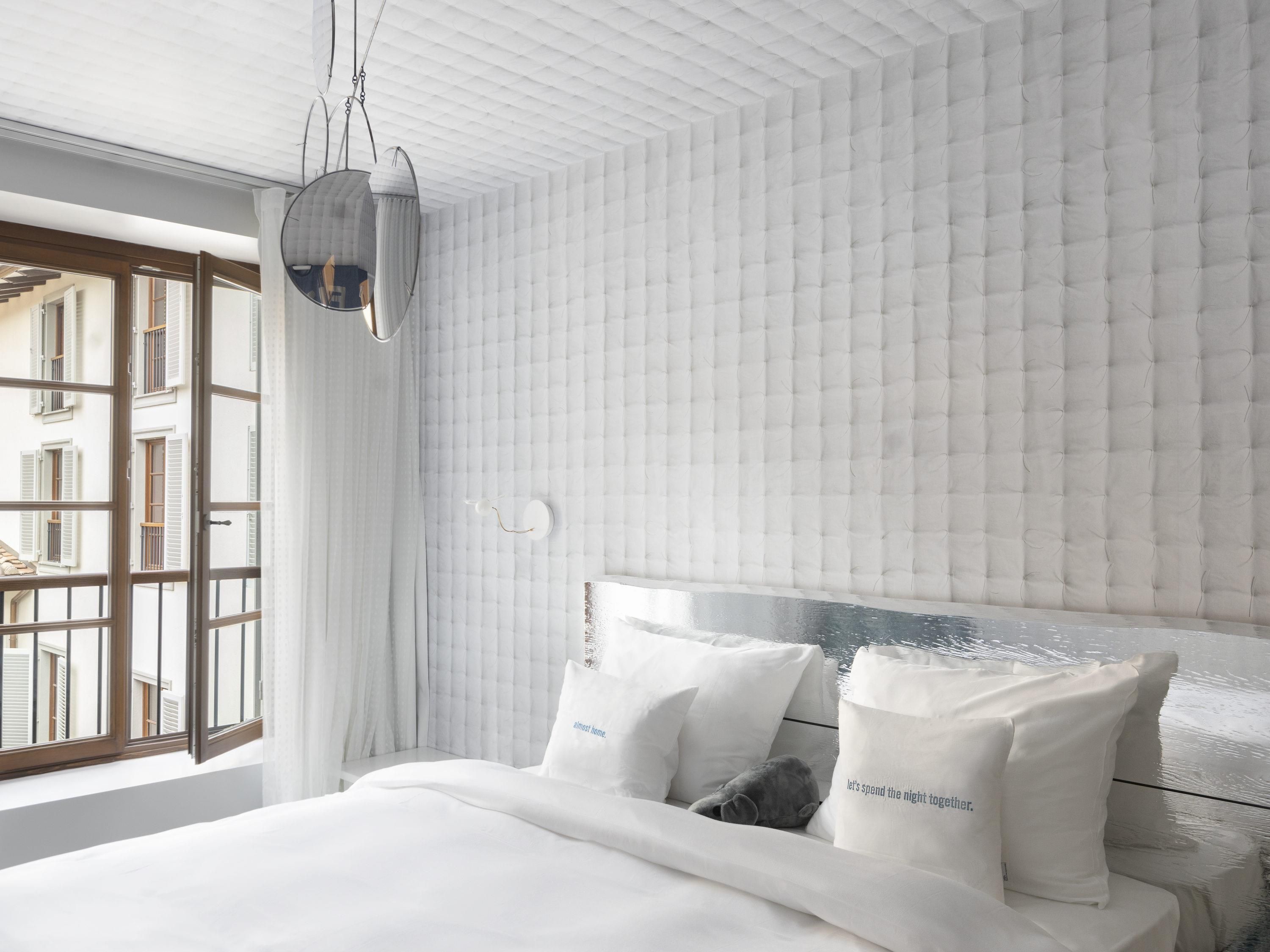 A modern bedroom with a large bed, white bedding, and a view of buildings through open windows.