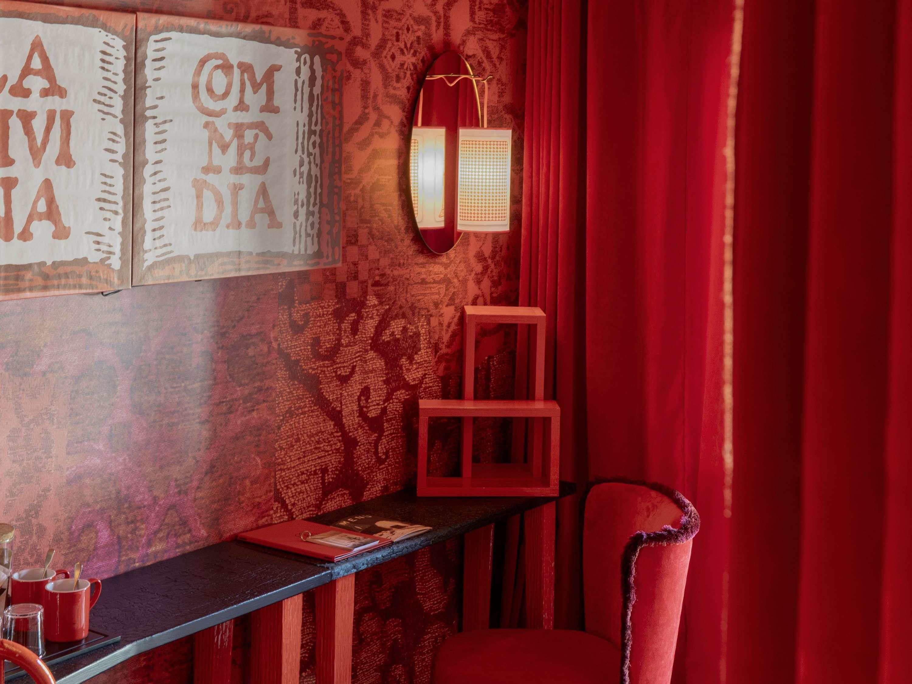 A room with red curtains, a red chair, a table with a red lamp, and wall art with text.
