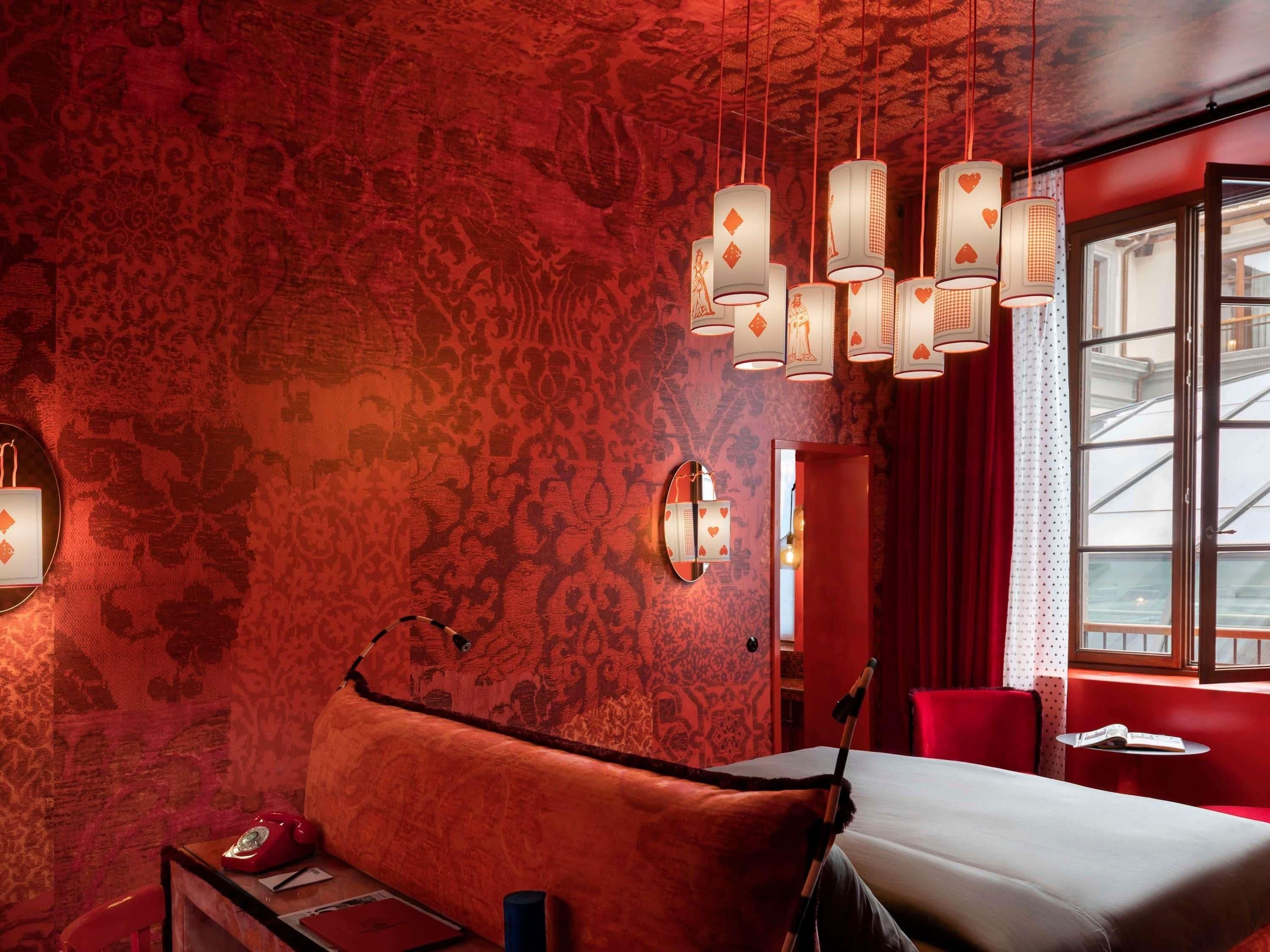A hotel room with a red patterned wall, a bed, a red armchair, a small table with a book, and a window with a view.