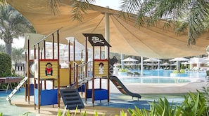 A playground with slides and climbing structures, a pool area with lounge chairs, and palm trees.