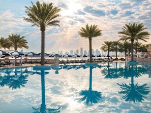 A pool with palm trees and lounge chairs, a city skyline in the background, and a clear sky.