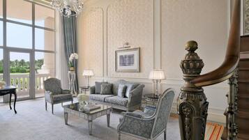 A spacious living room with a grand chandelier, elegant furniture, and a large window offering a view of greenery.