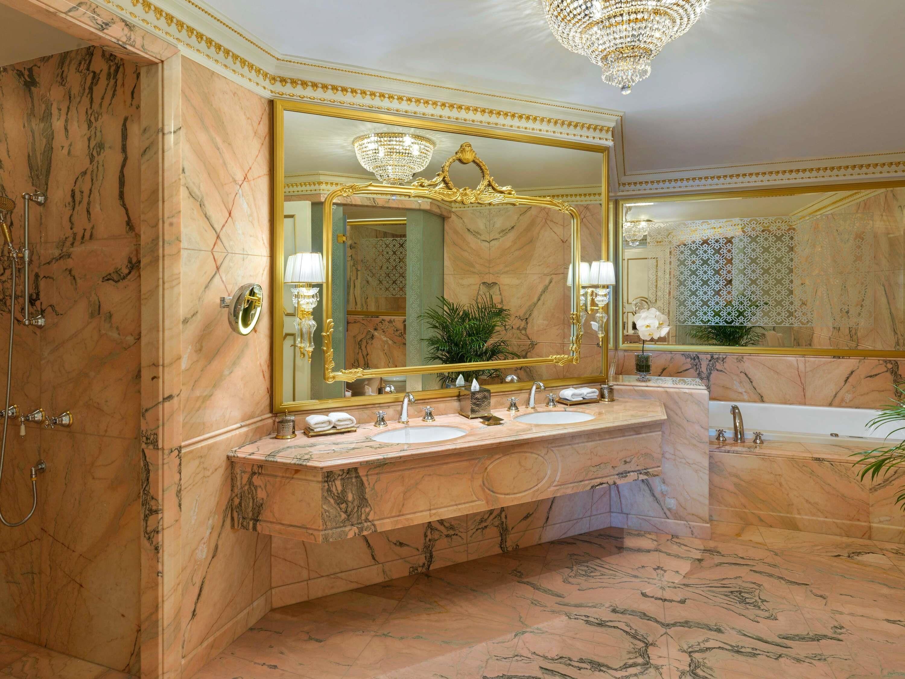 A luxurious bathroom with marble walls, a large mirror, a chandelier, and a bathtub.