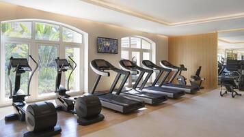 A gym with treadmills, elliptical machines, and a stationary bike, large windows with a view of greenery, and a wooden wall.